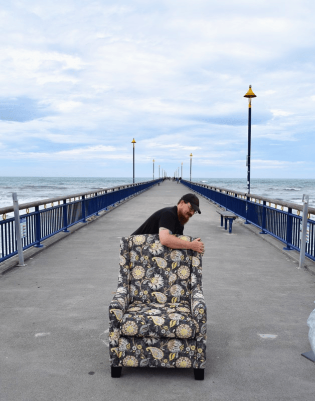 A man with a beard and black cap is playfully pushing a floral armchair on a pier, with the ocean in the background and cloudy skies.