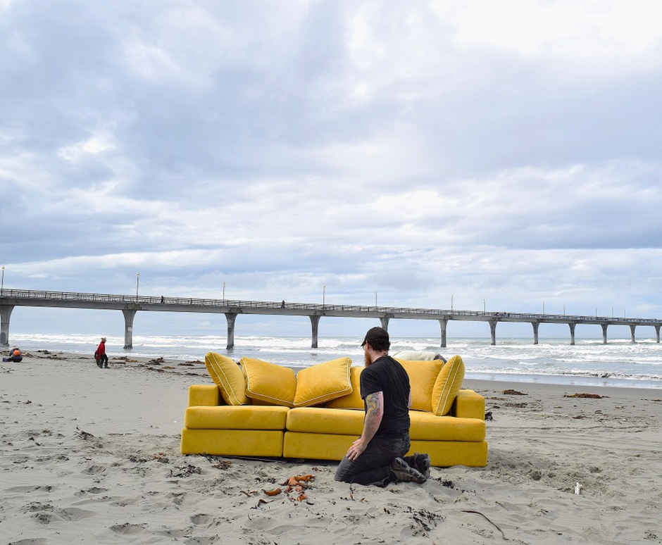 A person kneeling beside a bright yellow couch on a sandy beach, with a pier in the background and cloudy skies.