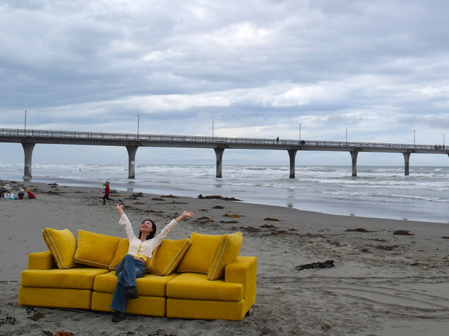 A woman sitting on a yellow couch on the beach with arms raised in joy, a pier visible in the background under a cloudy sky.