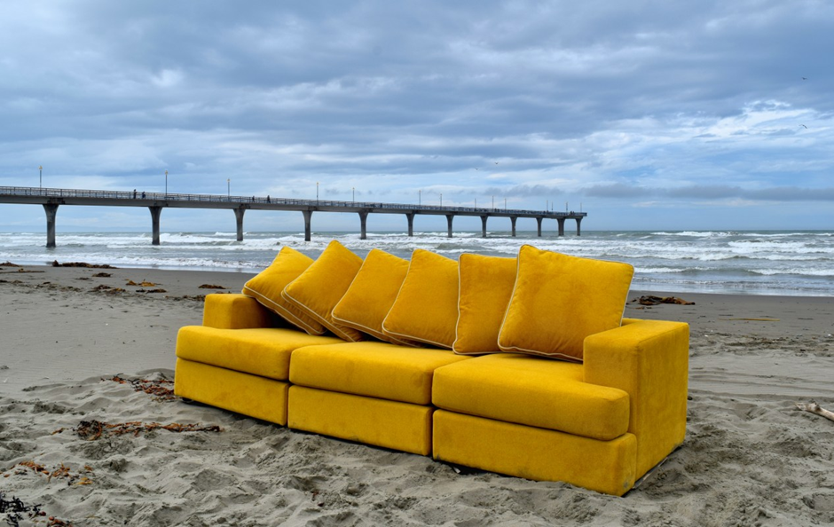 A vibrant yellow sofa with plush cushions is positioned on a sandy beach, with a pier extending into the ocean in the background under a cloudy sky.